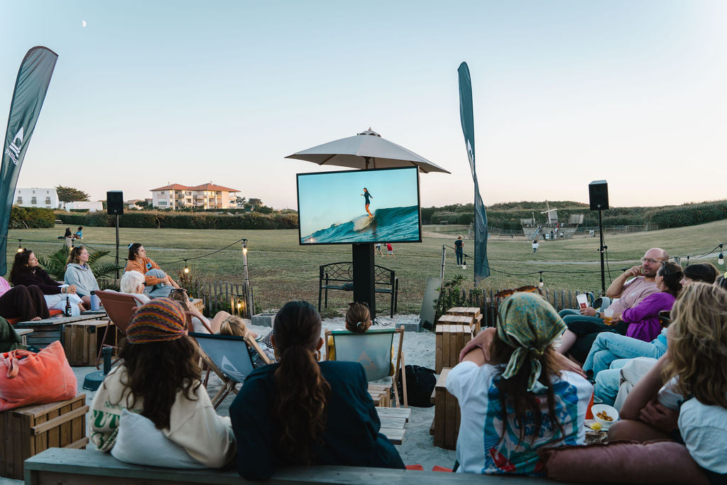 People sat around on deck chairs as the sun starts to set, watching a surf film on a large outdoor screen