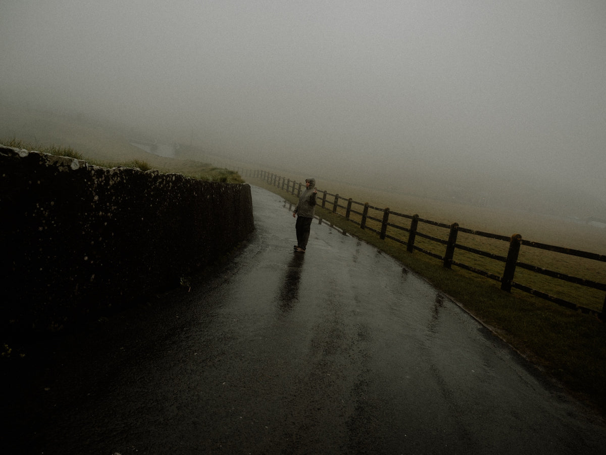 A person walks along a wet paved path in a foggy outdoor setting, bordered by a dark stone wall on the left and a wooden fence on the right.