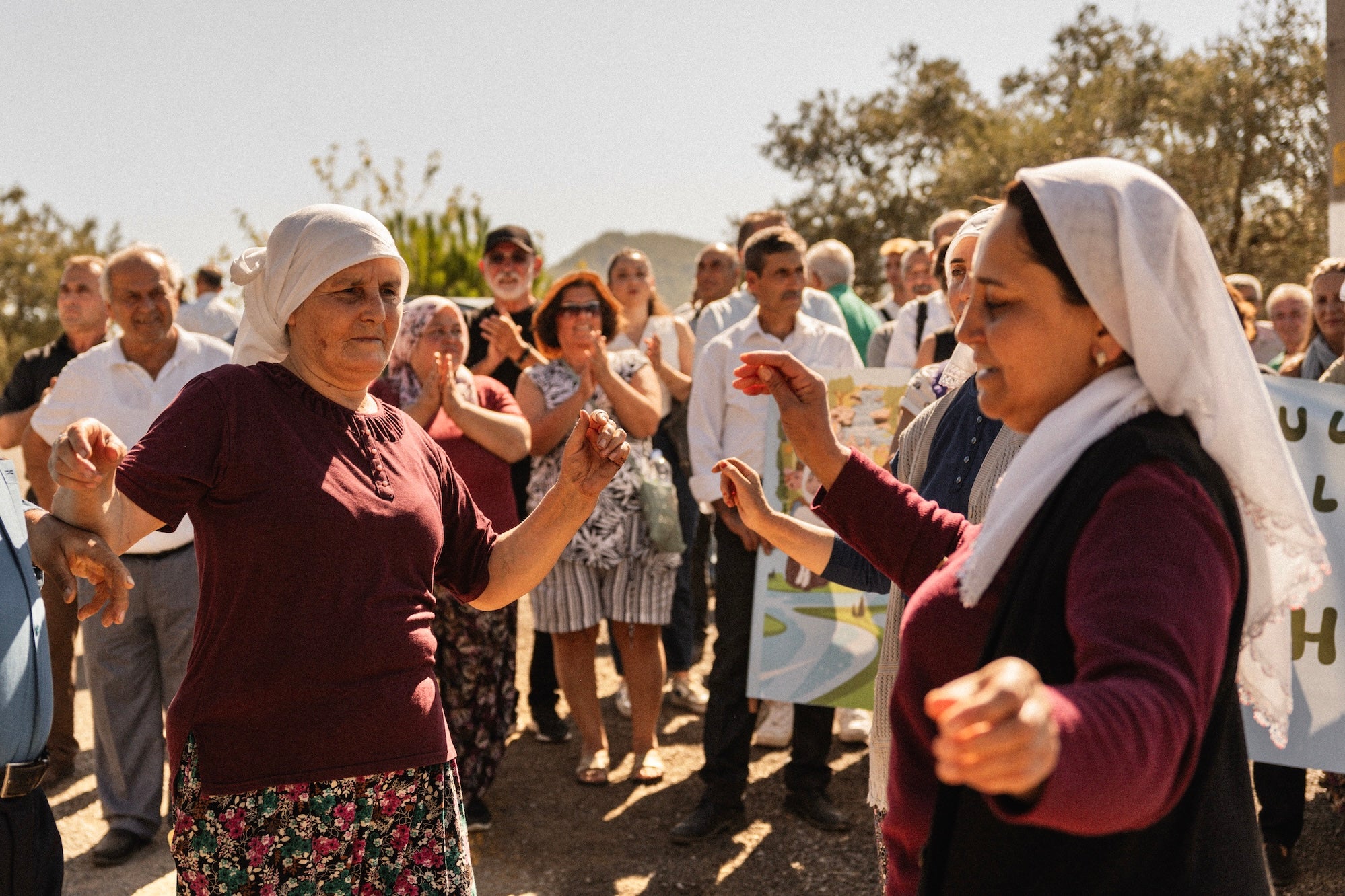Two women in headscarves dancing outdoors with a crowd in the background