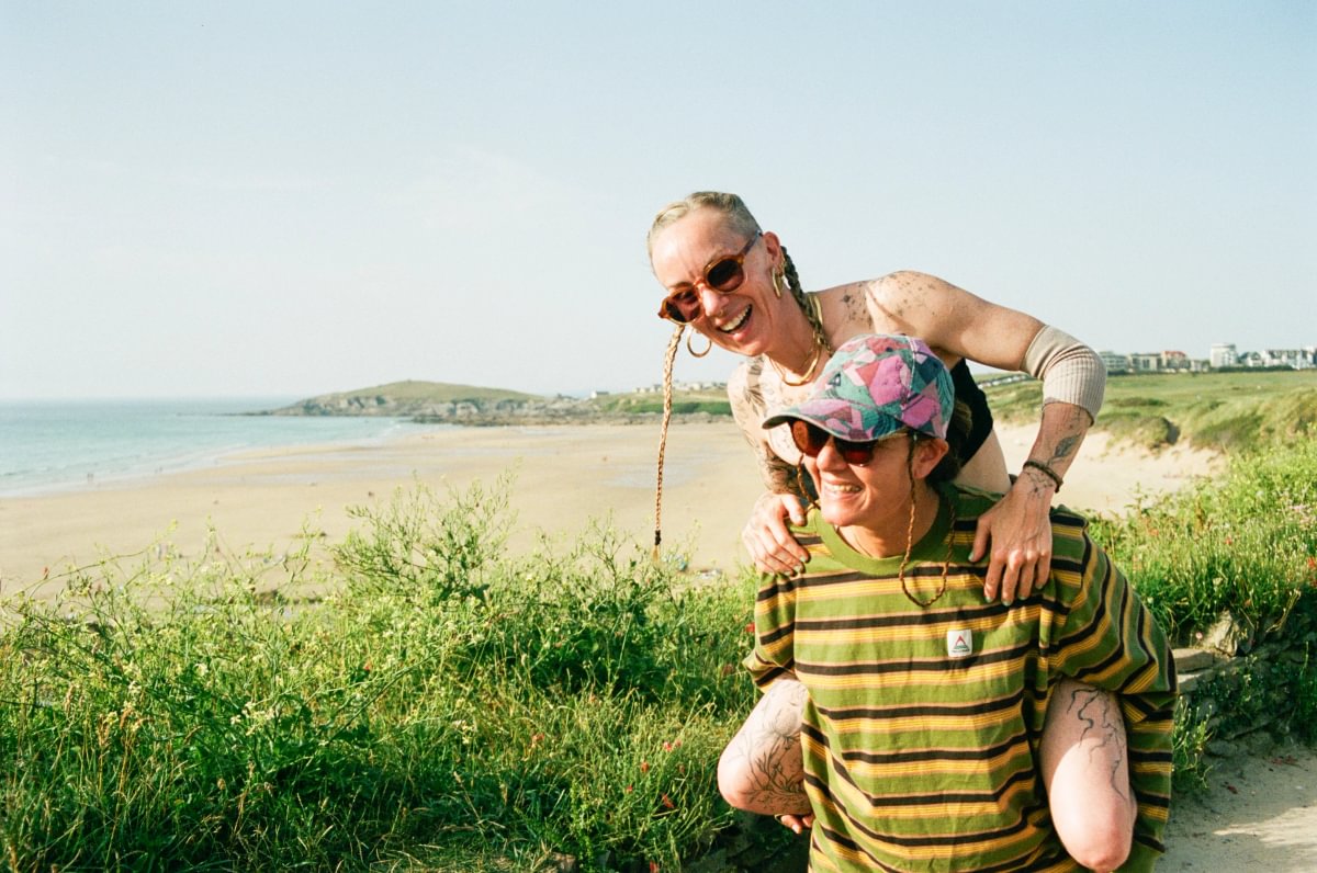 Two people outdoors on a beach dune with grass, sand, and water in the background. The person in front wears a green and yellow striped shirt and a colorful floral cap, while the person behind wears a beige sleeveless top and sunglasses.